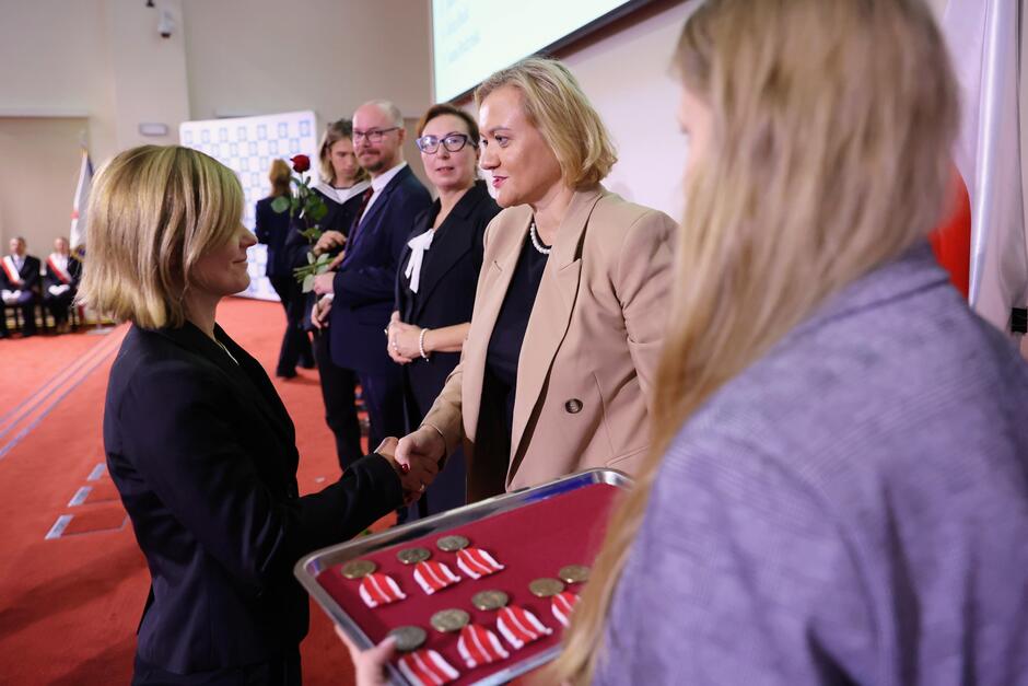 Zdjęcie przedstawia uroczystą ceremonię wręczania odznaczeń, gdzie jedna z kobiet, uśmiechnięta, odbiera medal od drugiej kobiety w jasnej marynarce. Na pierwszym planie widoczna jest taca z medalami, a w tle osoby trzymające róże, co sugeruje formalny charakter wydarzenia.