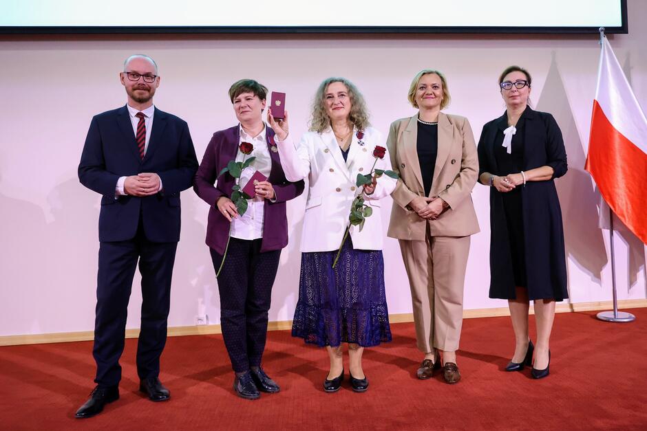 Zdjęcie przedstawia grupę osób podczas oficjalnej ceremonii, na której dwie kobiety otrzymują odznaczenia, trzymając w dłoniach czerwone róże i medale. Uczestnicy stoją na czerwonym dywanie, a w tle widać polską flagę, co sugeruje, że jest to wydarzenie o charakterze państwowym.