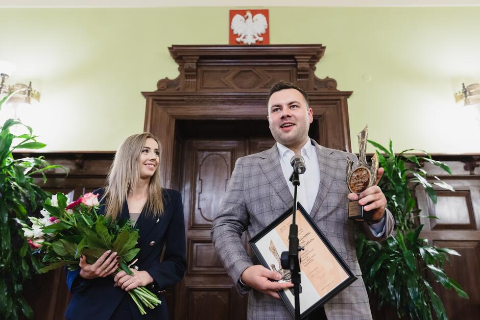 na zdjęciu młody mężczyzna trzyma nagrodę i mówi do mikrofonu, obok stoi młoda kobieta, patrzy na niego