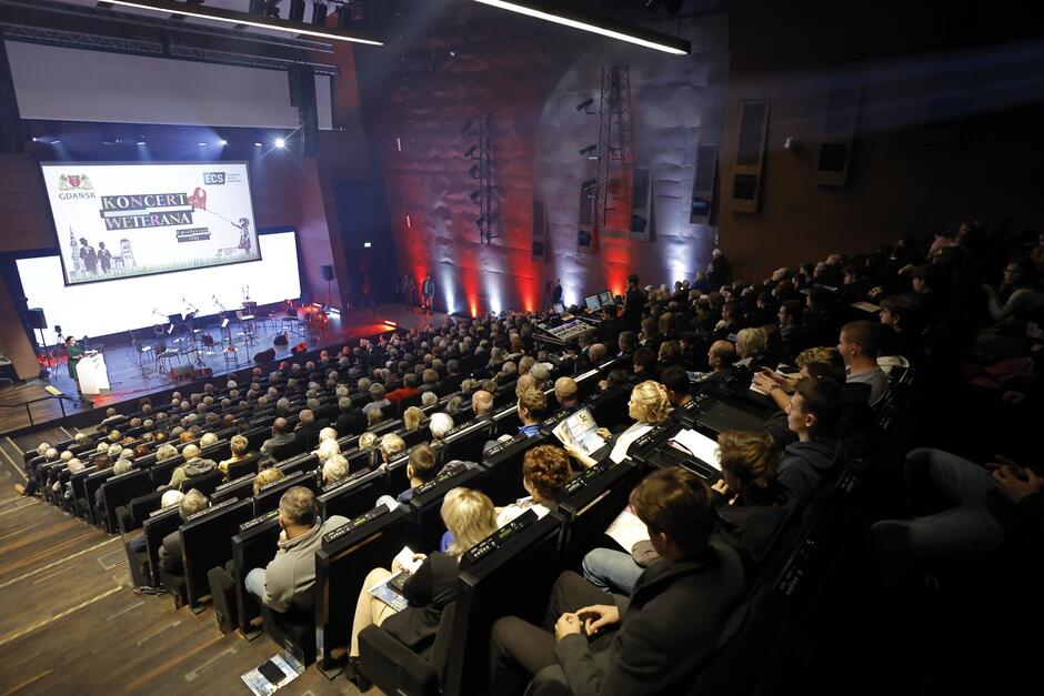 Sala koncertowa, na scenie przemawia kobieta. Na ekranie w tle napis "Koncert Weterana".