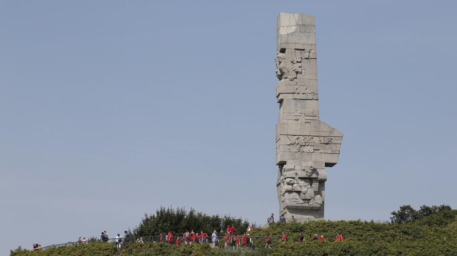 Na zdjęciu widoczny jest Pomnik Obrońców Wybrzeża znajdujący się na Westerplatte w Gdańsku. Pomnik został wzniesiony ku czci polskich żołnierzy, którzy bronili Westerplatte podczas II wojny światowej. Monument wykonany jest z szarego kamienia i ma formę wysokiej, geometrycznej kolumny ozdobionej reliefami przedstawiającymi sceny z walki oraz symbole związane z obroną. W dolnej części pomnika znajdują się ludzie, prawdopodobnie turyści, co pozwala ocenić skalę monumentu – jest on bardzo duży i imponujący. W tle widoczne jest jasne, bezchmurne niebo.