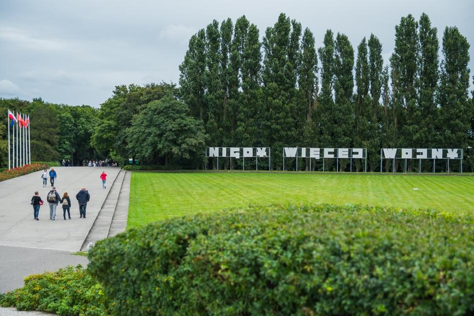 Zdjęcie przedstawia fragment terenu na Westerplatte w Gdańsku, gdzie znajduje się historyczne miejsce związane z wybuchem II wojny światowej. Na zdjęciu widać szeroką aleję, po której spacerują ludzie, a po lewej stronie stoi kilka flag na masztach, w tym flaga Polski oraz Unii Europejskiej. Po prawej stronie, na trawniku, znajduje się duży napis w języku polskim: „NIGDY WIĘCEJ WOJNY”, umieszczony na metalowych podporach. W tle widoczny jest pas wysokich drzew, które stanowią naturalną granicę dla tego terenu. Atmosfera zdjęcia jest spokojna, a teren wygląda na zadbany.