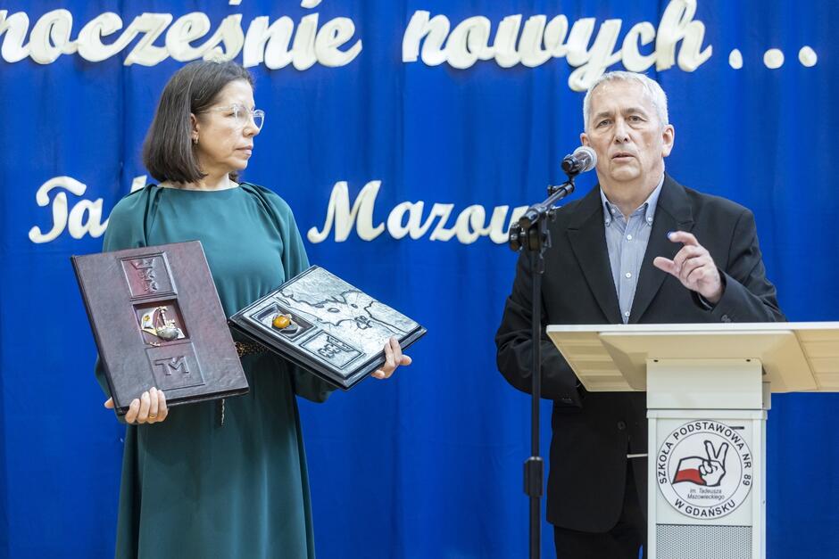 Na zdjęciu znajduje się dwóch dorosłych ludzi na tle niebieskiego baneru z białym napisem. Kobieta po lewej stronie, ubrana w zieloną sukienkę i okulary, trzyma w rękach dwa duże, ozdobne albumy lub księgi. Wygląda na to, że są to przedmioty o dużym znaczeniu, być może symboliczne lub pamiątkowe. Mężczyzna po prawej stronie, ubrany w czarną marynarkę i koszulę, stoi za mównicą z logo Szkoły Podstawowej nr 8 im. Tadeusza Mazowieckiego w Gdańsku. Wygłasza przemówienie, mówiąc do mikrofonu i wykonując gest ręką. Cała scena sugeruje uroczystość szkolną, być może związana z wręczeniem nagród lub pamiątek.