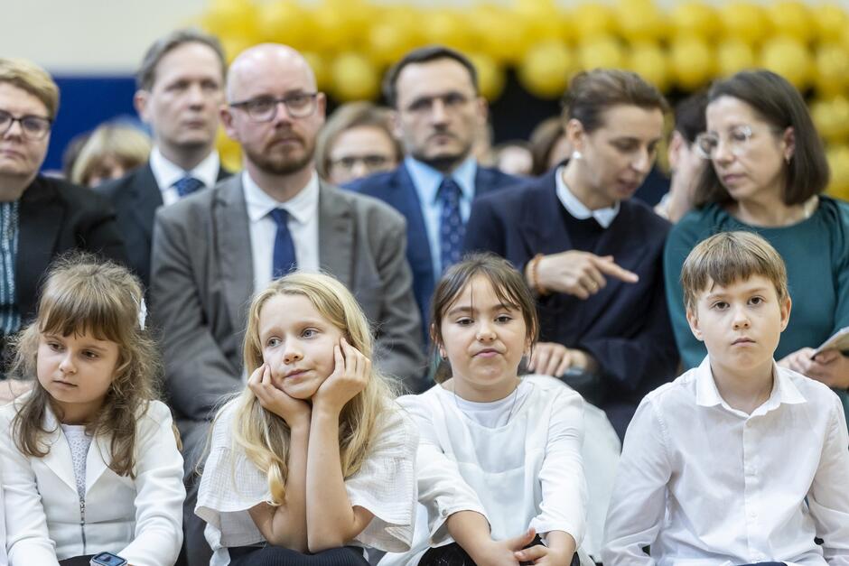 Na zdjęciu widzimy grupę dzieci siedzących w pierwszym rzędzie podczas uroczystości. Cztery dzieci są ubrane elegancko w białe bluzki, co może sugerować, że biorą udział w ważnym wydarzeniu, takim jak akademia szkolna. Dziewczynki z lewej strony wyglądają na lekko znudzone lub zamyślone. Jedna z nich podpiera głowę dłońmi, co wskazuje na to, że wydarzenie może trwać już jakiś czas. Chłopiec po prawej stronie ma poważny wyraz twarzy. W tle za dziećmi siedzą dorośli, również elegancko ubrani, w garniturach i formalnych strojach, co dodatkowo podkreśla uroczysty charakter wydarzenia. Widać, że dwie kobiety rozmawiają między sobą, jedna wskazuje palcem na coś przed sobą. Tło dekorowane jest żółtymi balonami, co sugeruje, że jest to specjalna okazja, być może rocznicowa akademia lub zakończenie roku szkolnego.