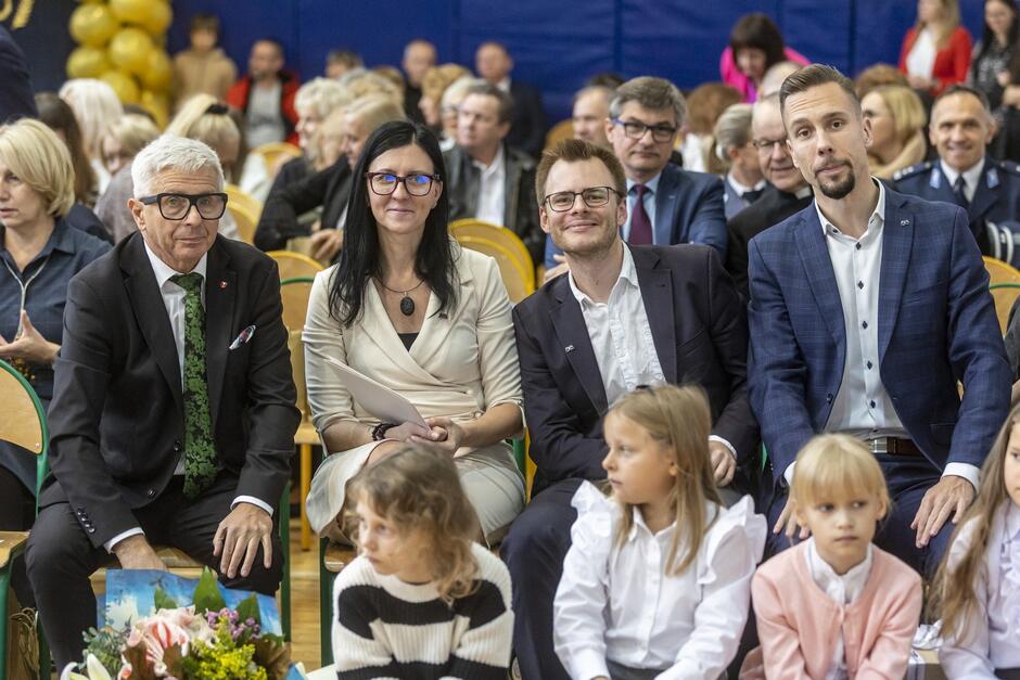 Na zdjęciu widzimy grupę elegancko ubranych dorosłych siedzących w pierwszym rzędzie na uroczystości, najprawdopodobniej szkolnej lub rocznicowej. Na pierwszym planie siedzi czwórka dzieci, które prawdopodobnie również uczestniczą w wydarzeniu. Dzieci są ubrane w jasne, odświętne stroje, a jedna z dziewczynek trzyma bukiet kwiatów, co sugeruje, że mogą one pełnić jakąś rolę w ceremonii. Wśród dorosłych na uwagę zwraca mężczyzna po lewej stronie, ubrany w elegancki garnitur z zielonym krawatem, obok niego siedzi kobieta w kremowej sukience, a obok niej dwóch młodszych mężczyzn w ciemnych marynarkach. Cała grupa wygląda na uśmiechniętą i zrelaksowaną, co sugeruje, że jest to miła, uroczysta okazja. W tle widać innych uczestników wydarzenia, siedzących na krzesłach w sali gimnastycznej, a dekoracje z żółtych balonów nawiązują do świątecznego charakteru uroczystości.