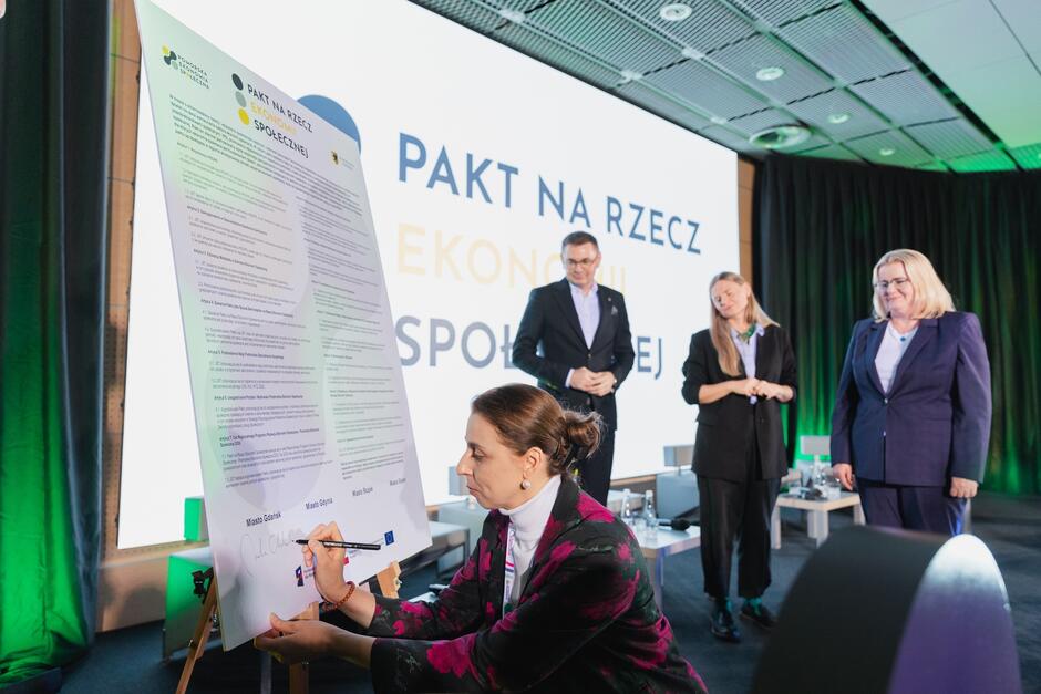 Kobieta podpisuje się na tablicy z treścią porozumienia na sali konferencyjnej.