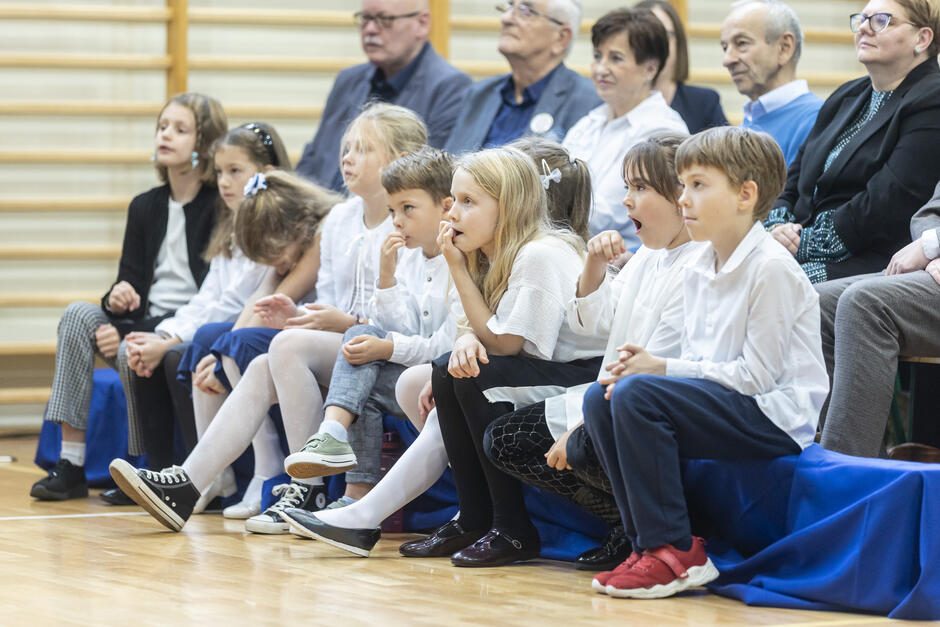 grupa odświętnie ubranych dzieci siedzi na drewnianej ławce w sali gimnastycznej, za nimi widoczna grupa dorosłych