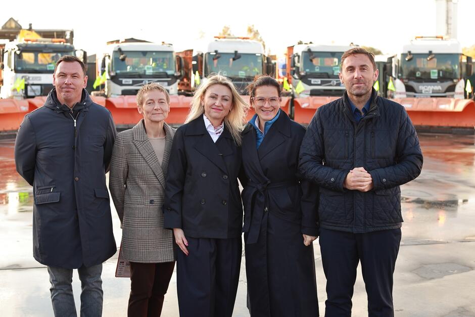 Na zdjęciu widoczna jest grupa pięciu osób stojących na tle rzędu pługów śnieżnych. Uśmiechają się do kamery, a w tle znajdują się pojazdy przygotowane do zimowego utrzymania dróg, co sugeruje, że jest to oficjalne spotkanie związane z przygotowaniami do sezonu zimowego.