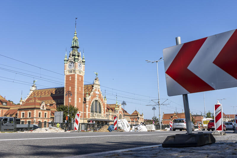 Na zdjęciu widać zabytkowy budynek dworca kolejowego z charakterystyczną wieżą zegarową, który znajduje się w Gdańsku. Na pierwszym planie widoczne są znaki drogowe informujące o prowadzonych pracach budowlanych, co sugeruje remont lub modernizację infrastruktury drogowej przed dworcem.