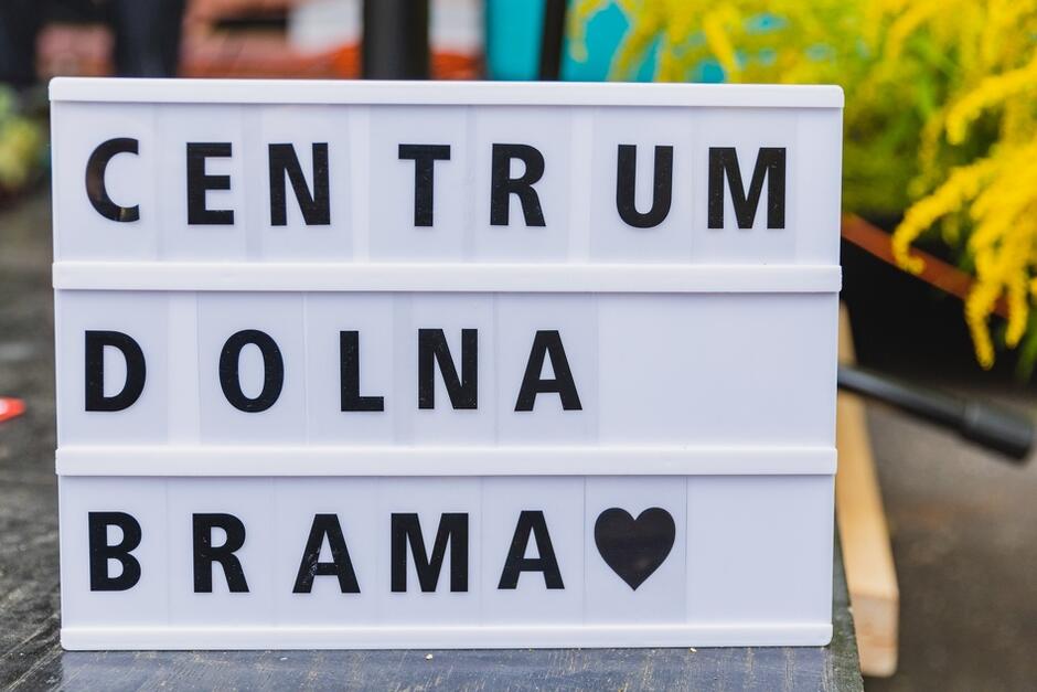 Zdjęcie przedstawia tablicę z napisem „CENTRUM DOLNA BRAMA” ułożonym z plastikowych liter na białym tle, a na dole widnieje symbol serca. W tle można dostrzec kolorowe elementy dekoracyjne, co sugeruje, że zdjęcie może pochodzić z jakiegoś wydarzenia związanego z tym miejscem.