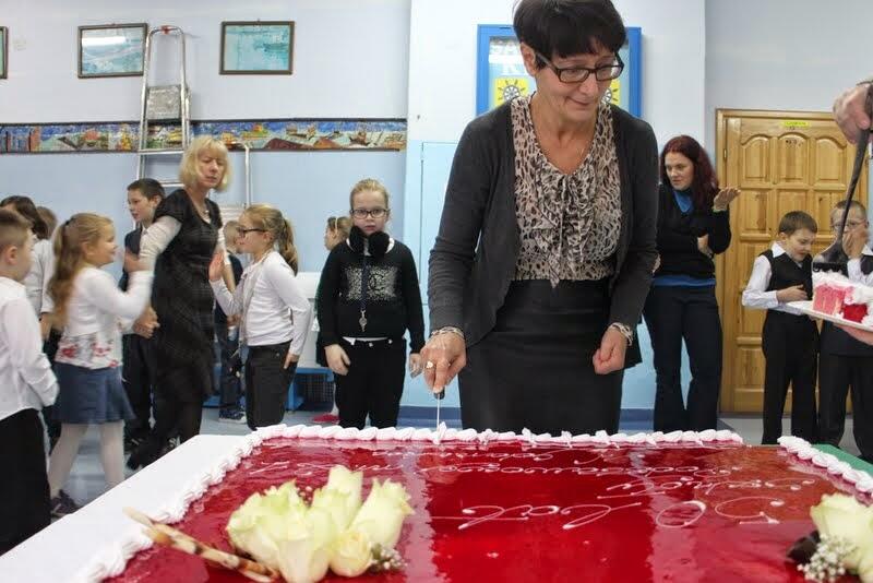 Zdjęcie przedstawia uroczyste krojenie dużego tortu podczas szkolnej imprezy. Na pierwszym planie widzimy kobietę, najprawdopodobniej nauczycielkę, która trzyma nóż i przygotowuje się do krojenia tortu. Tort jest pokryty czerwonym lukrem, ozdobiony białymi różami oraz napisami, choć napisy są częściowo niewyraźne na zdjęciu. W tle widać grupę dzieci, które stoją w kolejce, najprawdopodobniej czekając na kawałki tortu. Niektóre dzieci są ubrane elegancko, co sugeruje, że to specjalna okazja, być może związana z jakimś szkolnym świętem lub zakończeniem projektu. Sala ozdobiona jest w sposób klasyczny, z obrazkami na ścianach, a z lewej strony widoczna jest drabina, co może sugerować przygotowania do dekorowania lub sprzątania po wydarzeniu.  Atmosfera na zdjęciu jest radosna, a dzieci wydają się podekscytowane, czekając na swoją kolej.