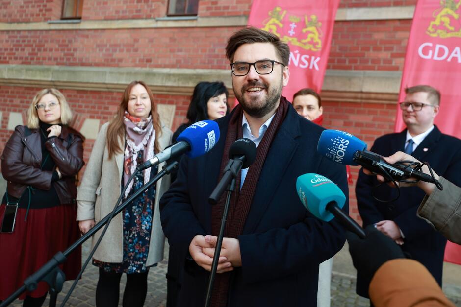 Na zdjęciu znajduje się mężczyzna w okularach, który uśmiecha się i przemawia do mikrofonu podczas konferencji prasowej. W tle stoi grupa kobiet oraz mężczyzna, a za nimi widoczne są czerwone banery z napisem Gdańsk  i herbem miasta