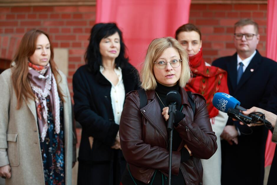 Na zdjęciu widzimy kobietę w okularach i skórzanej kurtce, która przemawia do mikrofonu podczas konferencji prasowej. Za nią stoi kilka osób, w tym kobieta w czerwonym szalu oraz mężczyzna w garniturze, którzy słuchają jej wypowiedzi