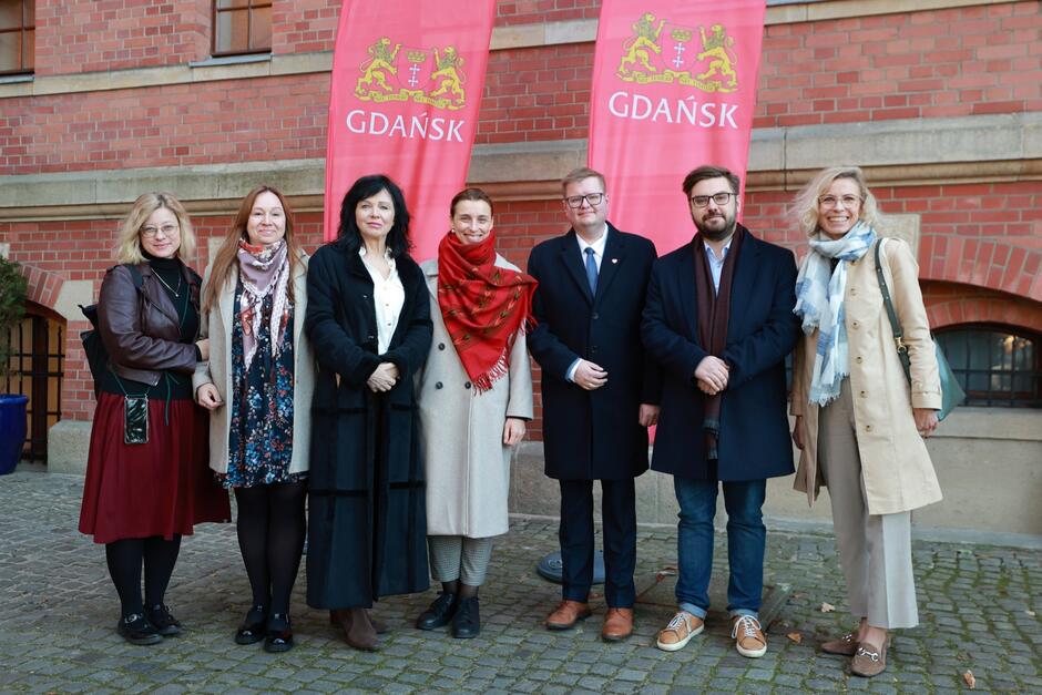 Na zdjęciu znajduje się grupa siedmiu osób pozujących do wspólnego zdjęcia na tle dwóch czerwonych banerów z napisem "Gdańsk" i herbem miasta. Wszyscy są ciepło ubrani, a wśród nich znajdują się zarówno kobiety, jak i mężczyźni, stojący w rzędzie na tle ceglanego budynku