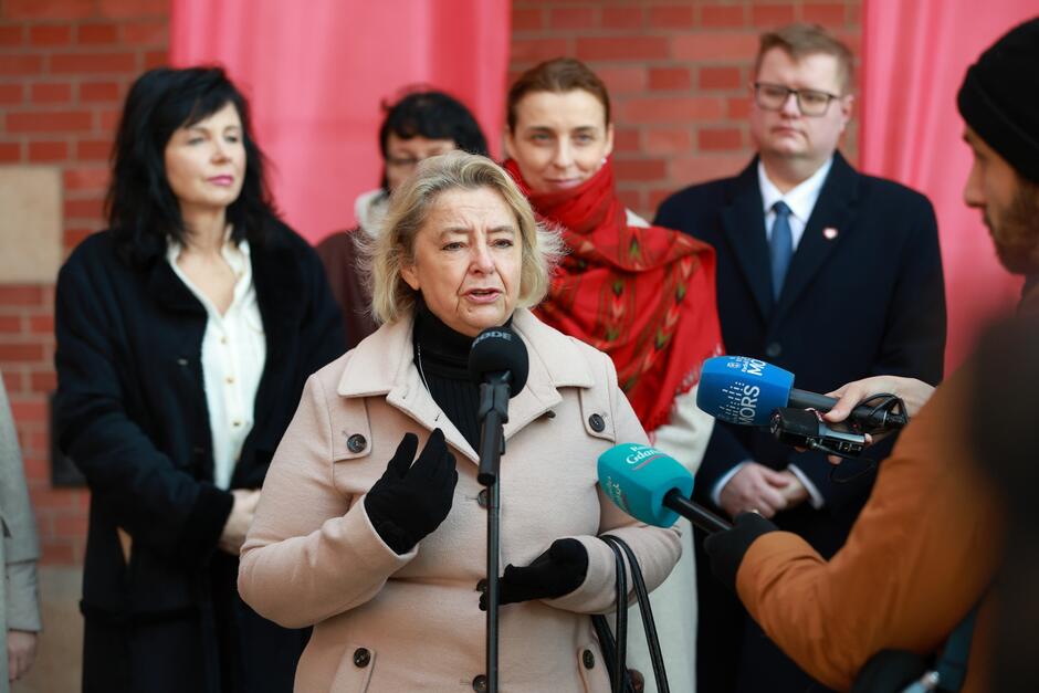 Na zdjęciu widzimy kobietę w jasnym płaszczu, która przemawia do mikrofonu podczas konferencji prasowej. W tle stoi grupa osób, w tym mężczyzna w garniturze oraz kobieta z szalem, słuchających jej wystąpienia