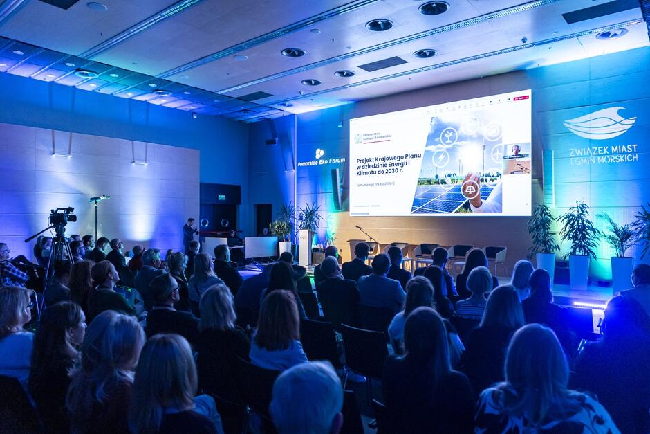 Sala konferencyjna, w której siedzą ludzie, na ekranie widać prezentację.