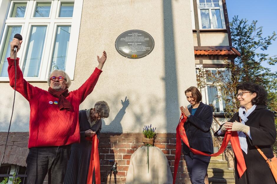 Na zdjęciu widzimy mężczyznę w czerwonym swetrze i okularach, który unosi ręce, przemawiając do mikrofonu podczas uroczystości odsłonięcia tablicy pamiątkowej na ścianie budynku. Obok niego stoją trzy kobiety, z których dwie trzymają wstęgę, a jedna przygotowuje się do jej przecięcia