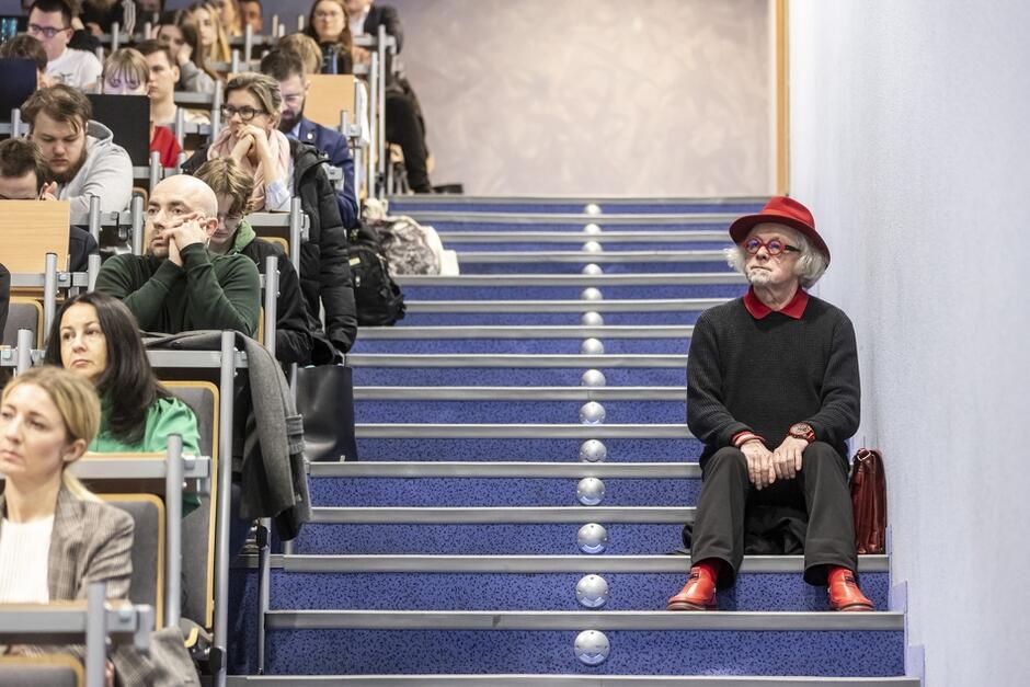 Na zdjęciu widzimy salę wykładową, gdzie siedzą ludzie słuchający prezentacji lub wykładu. Po prawej stronie, na schodach, siedzi starszy mężczyzna w czerwonym kapeluszu, czerwonych butach i okularach, wyróżniając się swoim oryginalnym strojem na tle innych uczestników