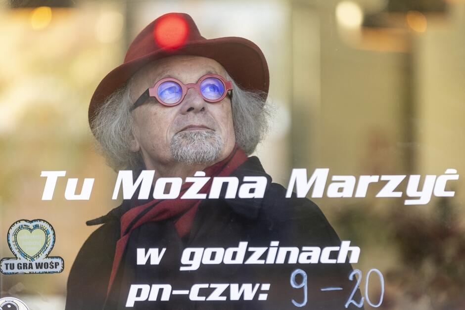 Na zdjęciu widzimy mężczyznę w czerwonym kapeluszu i czerwonych okularach, patrzącego przez szybę z napisem "Tu Można Marzyć". Napis informuje również o godzinach otwarcia miejsca: od poniedziałku do czwartku, od 9:00 do 20:00