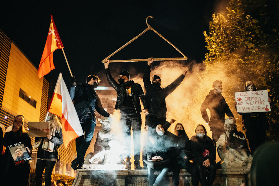 Na zdjęciu widzimy grupę protestujących, z których część stoi na pomniku, wznosząc ręce w geście sprzeciwu, trzymając flagi Polski oraz duży metalowy wieszak – symbol walki o prawa kobiet. Otoczeni są dymem, co podkreśla dramatyzm sceny, a ich twarze są częściowo zakryte maskami. Wśród zgromadzonych widać transparenty z hasłami wyrażającymi ich niezadowolenie wobec aktualnej sytuacji politycznej, co sugeruje, że zdjęcie przedstawia moment społecznego protestu.