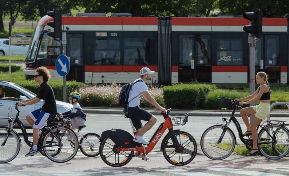 Na zdjęciu widzimy grupę osób na rowerach poruszających się po ścieżce rowerowej obok ulicy, na której znajduje się tramwaj. W tle widać zieleń oraz charakterystyczne oznakowanie drogowe, co sugeruje miejskie otoczenie i promowanie transportu ekologicznego.