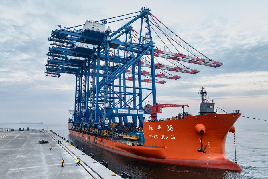 Na zdjęciu widoczny jest ogromny statek transportowy o nazwie Zhen Hua 36, należący do firmy ZPMC. Statek jest pomalowany na pomarańczowo i przewozi duże, niebieskie dźwigi portowe, które wystają wysoko ponad pokład. Dźwigi mają rozłożone ramiona, co nadaje im charakterystyczny wygląd przypominający ogromne żurawie portowe. Widać również pracowników w kamizelkach odblaskowych znajdujących się na nabrzeżu i przy statku, co wskazuje na operacje związane z rozładunkiem lub obsługą sprzętu.