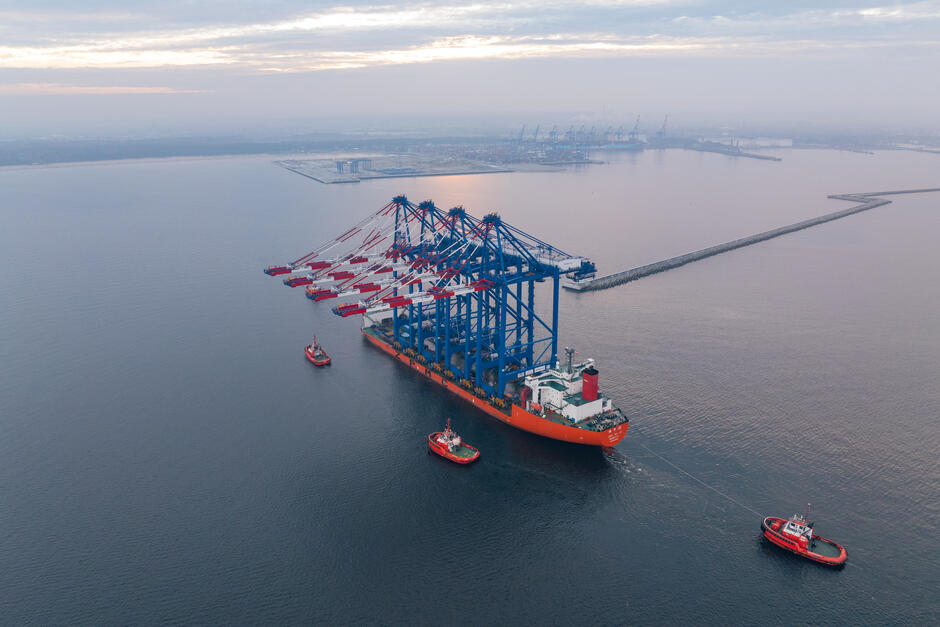 Na zdjęciu widać widok z lotu ptaka na ogromny statek transportowy Zhen Hua 36, który przewozi duże dźwigi portowe. Dźwigi te mają charakterystyczne niebieskie ramiona i czerwono-białe wysięgniki. Statek jest holowany przez trzy mniejsze holowniki w kierunku portu, co wskazuje na skomplikowaną manewrację przy wprowadzaniu go do nabrzeża. Otoczenie jest spokojne, z widocznym falochronem w oddali, a w tle można dostrzec zabudowania portowe i inne dźwigi, które rozciągają się na horyzoncie.