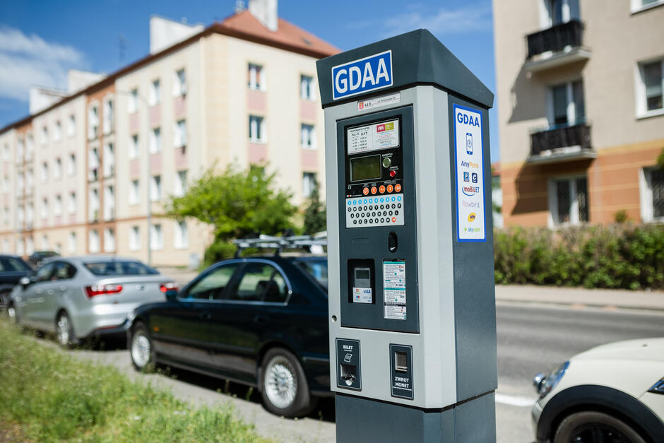 Na zdjęciu widać parkomat z oznaczeniem GDAA,  znajdujący się przy ulicy z zaparkowanymi samochodami na tle budynków mieszkalnych. Parkomat umożliwia płatności za pomocą różnych aplikacji, takich jak AnyPark i moBILET.