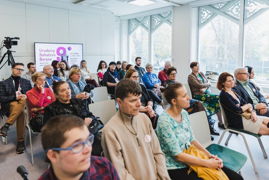 Na zdjęciu widzimy grupę osób siedzących na sali konferencyjnej, uważnie słuchających prezentacji lub wykładu. W tle znajduje się ekran z informacją o wydarzeniu zatytułowanym „Urodziny Bohatera Borysa”, sugerującym, że spotkanie ma charakter edukacyjny lub wspierający dla społeczności związanej z osobą Borysa.