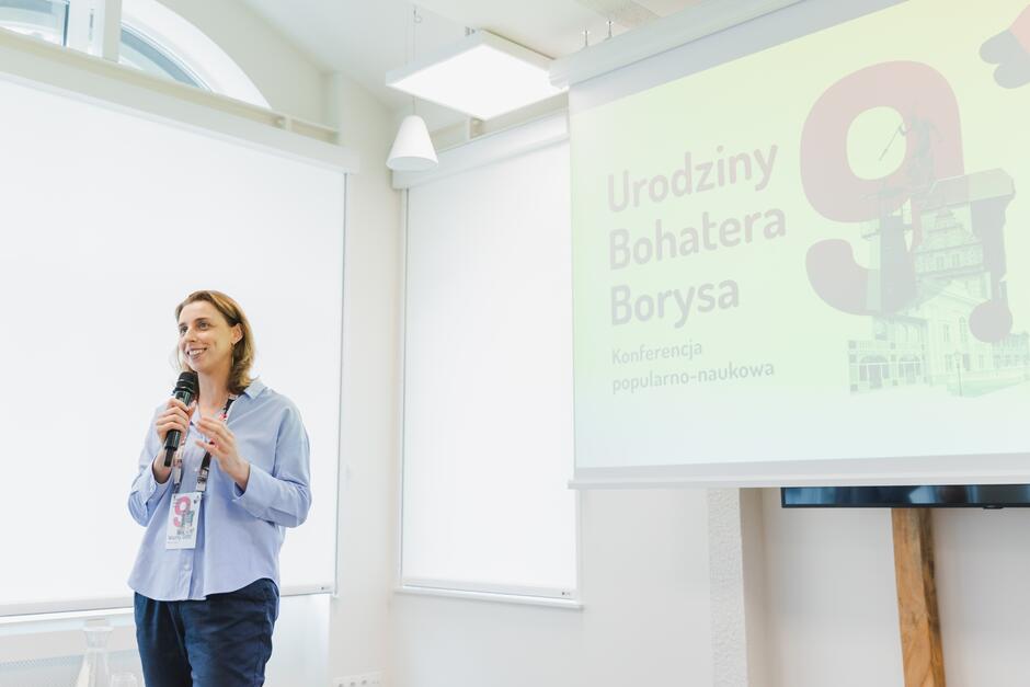 Na zdjęciu widzimy kobietę trzymającą mikrofon, która prowadzi prezentację na tle ekranu z napisem „Urodziny Bohatera Borysa – Konferencja popularno-naukowa.” Kobieta wygląda na uśmiechniętą i zaangażowaną, a jej identyfikator sugeruje, że jest częścią wydarzenia lub organizacji związanej z konferencją.