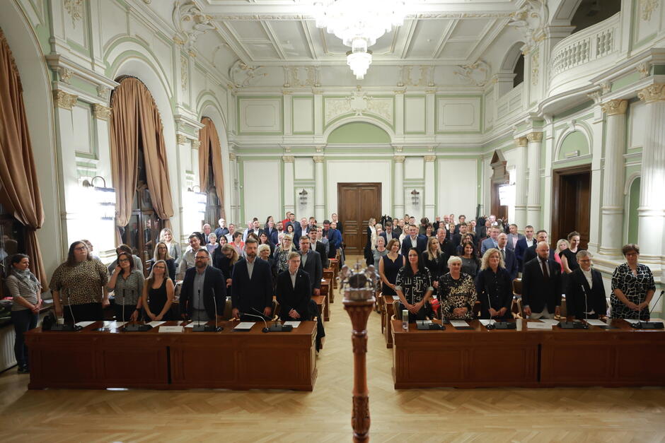 na zdjęciu ponad osiemdziesiąt osób, stoi w kilku rzędach, za masywnymi drewnianymi stołami, w historycznej sali ratuszowej