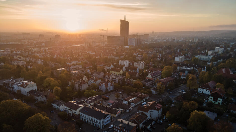 Zdjęcie przedstawia panoramę miasta o zachodzie słońca. Widać zabudowania mieszkalne, głównie niskie domy i kamienice, otoczone zielenią, co nadaje miastu przyjemny, spokojny charakter. W tle znajduje się wysoki, nowoczesny budynek, wybijający się ponad resztę zabudowy, oraz kilka innych wieżowców. Delikatne światło zachodzącego słońca nadaje całej scenerii ciepły, złocisty odcień, a unosząca się w powietrzu lekka mgła dodaje zdjęciu nastrojowego, melancholijnego klimatu.