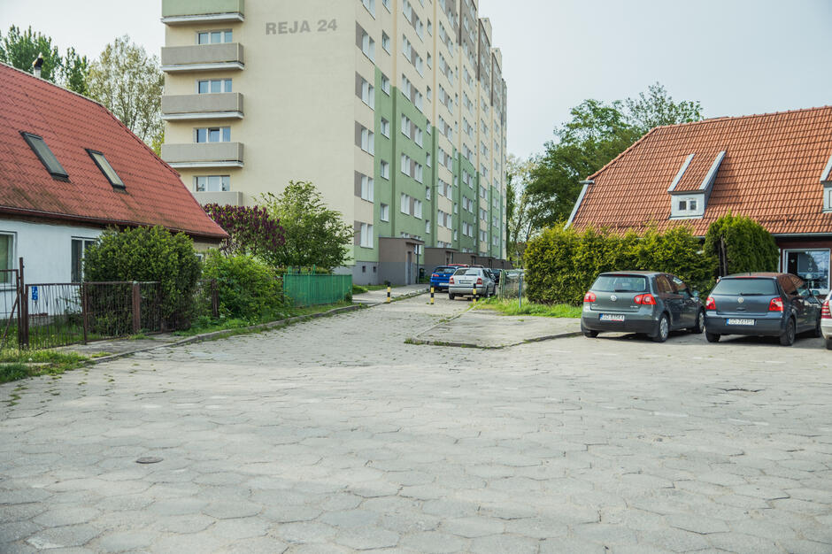 na zdjęciu po środku wysoki szary blok mieszkalny, po prawej i po lewej domki jednorodzinne, przed nimi nie wielki plac na którym stoją dwa samochody osobowe