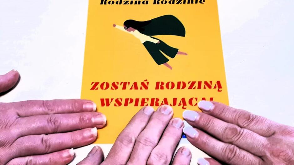 Grafika na białym tle leży kartka w żółtym kolorze z rysunkiem i napisem. Dłonie trzech osób dotykają kartki