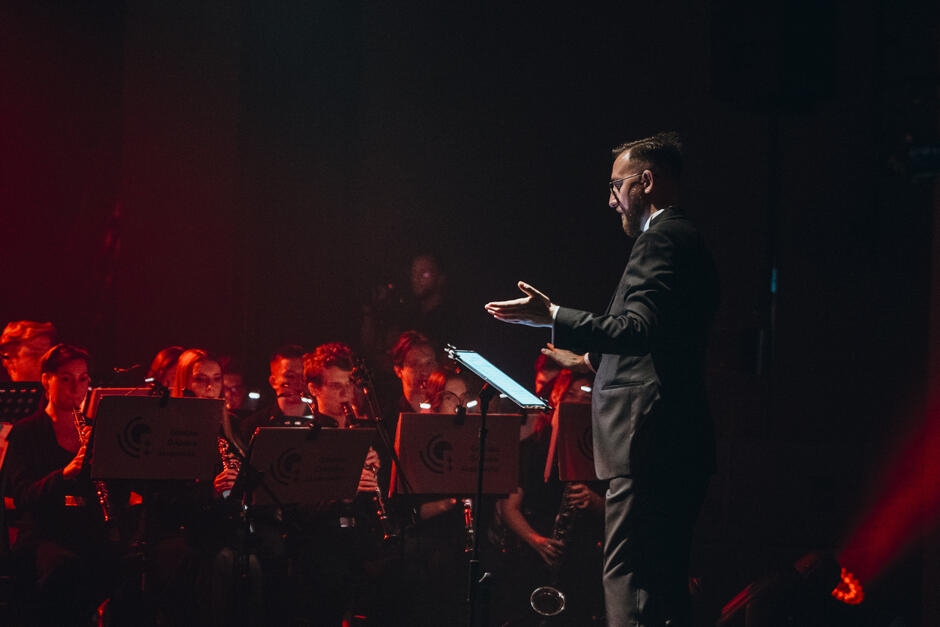 Na zdjęciu dyrygent prowadzi orkiestrę podczas występu, gestykulując z zaangażowaniem. W tle widać muzyków grających na instrumentach dętych, oświetlonych ciepłym, czerwonym światłem