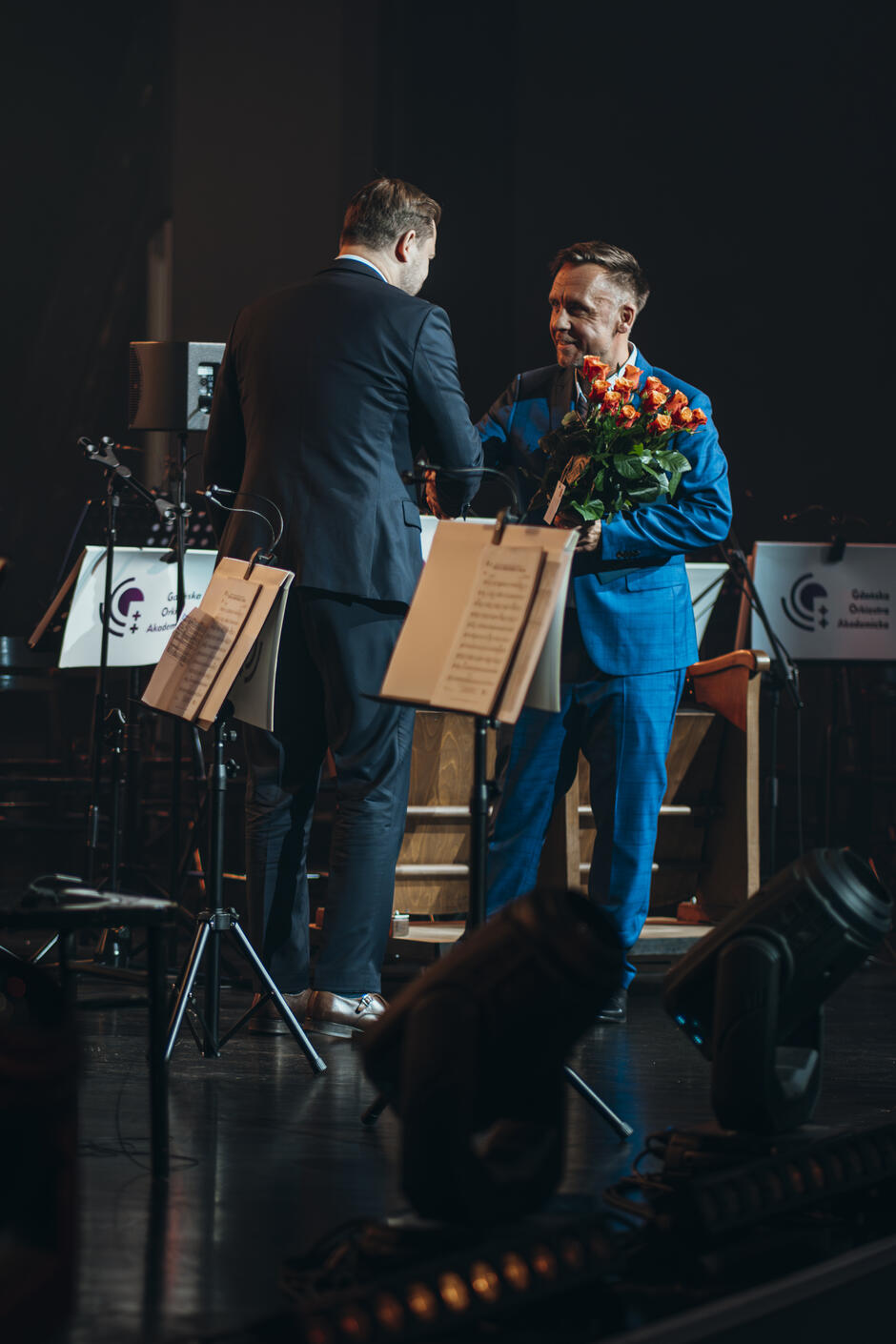 Na zdjęciu mężczyzna wręcza bukiet kwiatów drugiemu mężczyźnie na scenie. Obaj panowie są elegancko ubrani, a w tle widać pulpity z logo Gdańskiej Orkiestry Akademickiej