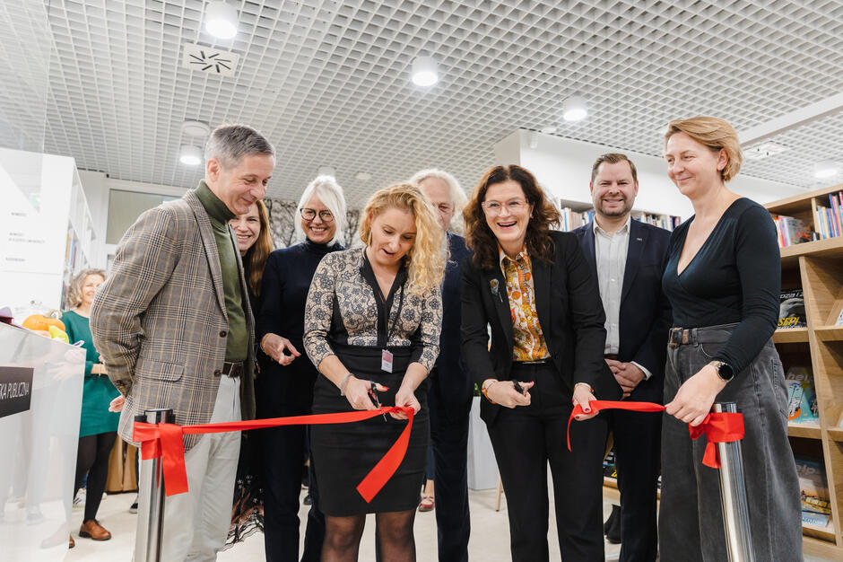 Na zdjęciu widzimy grupę uśmiechniętych osób podczas ceremonii przecięcia wstęgi w bibliotece, dwie kobiety w środku trzymają po parze nożyczek