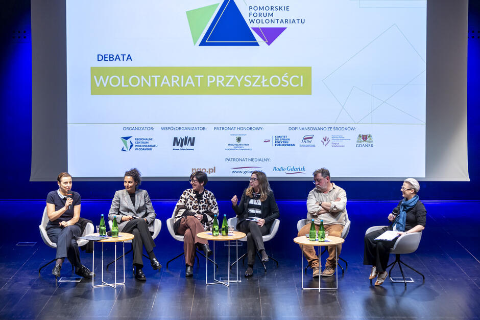 Na zdjęciu widzimy panel dyskusyjny podczas Pomorskiego Forum Wolontariatu, zatytułowany Wolontariat Przyszłości , z udziałem sześciu osób siedzących na scenie i prowadzących rozmowę. W tle wyświetlony jest ekran z logotypem wydarzenia oraz logotypami organizatorów i partnerów, co podkreśla formalny charakter spotkania.