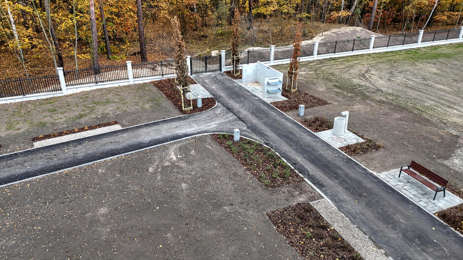 Zdjęcie przedstawia nowo wybudowaną część cmentarza, z wyraźnie wytyczonymi asfaltowymi ścieżkami i wydzielonymi miejscami na ławki oraz kosze na śmieci. W tle widać ogrodzenie z białymi słupkami i ciemnymi przęsłami, otoczone jesiennym lasem