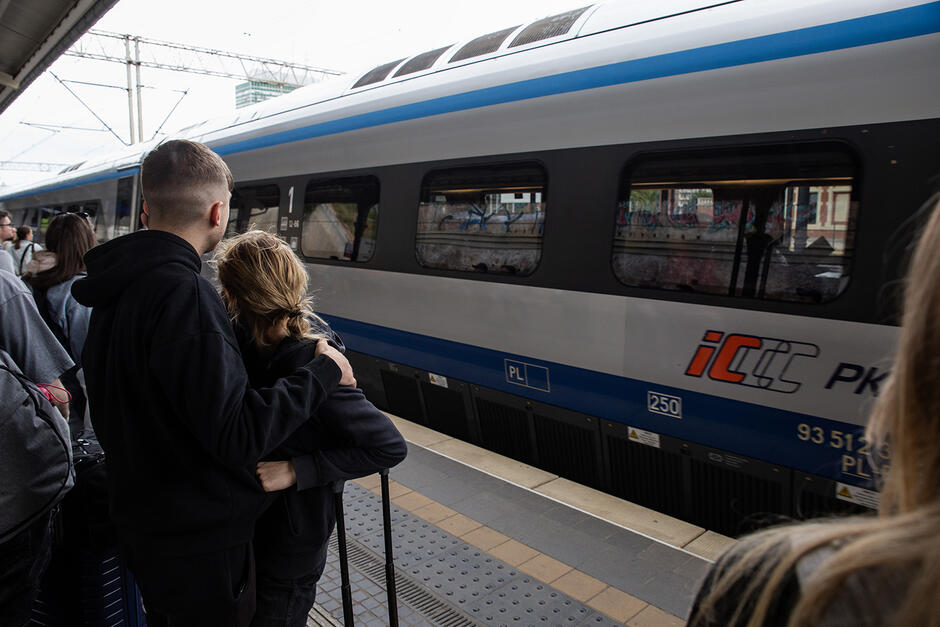 Na peronie kolejowym stoją ludzie, młody mężczyzna obejmuje młoda kobietę. Po torach jedzie pociąg.