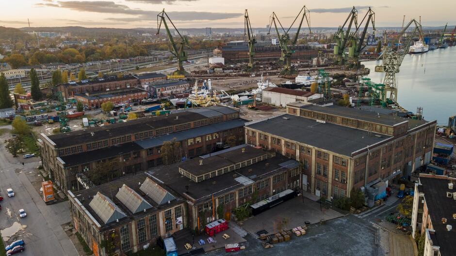 Na zdjęciu widzimy panoramę stoczni przemysłowej zlokalizowanej nad wodą. Na pierwszym planie znajdują się duże, historyczne hale fabryczne z cegły i betonu, o nieco zniszczonej elewacji, co wskazuje na długi czas ich użytkowania. Budynki mają prostą, industrialną architekturę i są rozmieszczone w sposób charakterystyczny dla terenów przemysłowych. W dalszej części obrazu widać duże żurawie stoczniowe, charakterystyczne dla tego typu terenów. Stoją one w rzędzie, górując nad pozostałymi strukturami i nadając tej przestrzeni typowy portowy krajobraz. Widać również statki i inne elementy infrastruktury stoczniowej, świadczące o aktywnej działalności przemysłowej.  W tle widać pas zieleni i dalsze zabudowania miejskie, które oddzielają stocznię od reszty miasta. Cała scena jest skąpana w ciepłym, wieczornym świetle, co nadaje jej spokojny i nostalgiczny charakter. Zdjęcie ukazuje przemysłowy urok tego miejsca oraz historyczne znaczenie, które stocznia odgrywała przez lata w życiu lokalnej społeczności.