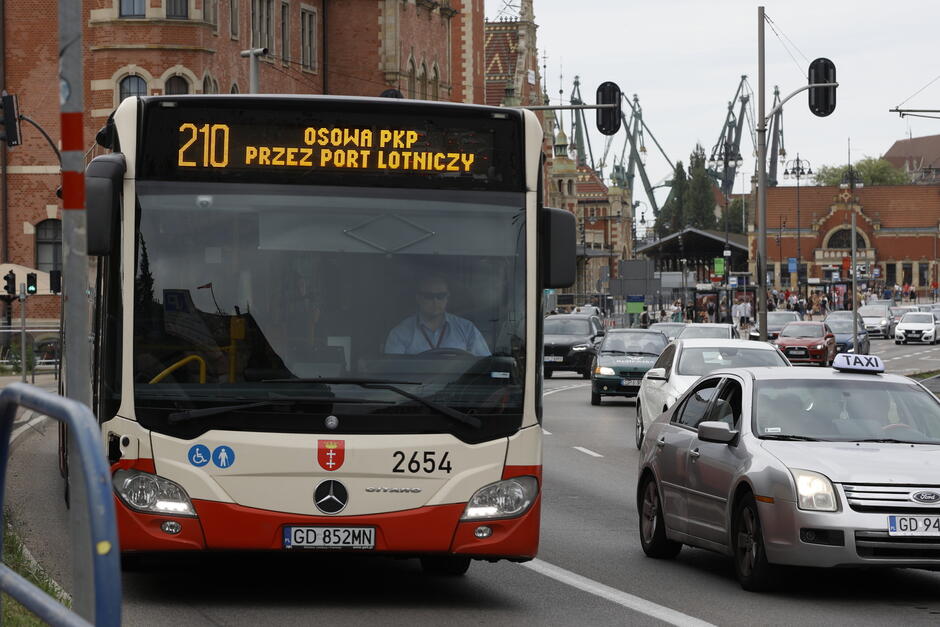 na zdjęciu autobus miejski z numerem dwieście dziesięć, widać prowadzącego go kierowcę, autobus jedzie dość zatłoczoną ulicą widać na pozostałych pasach jezdni sporo samochodów