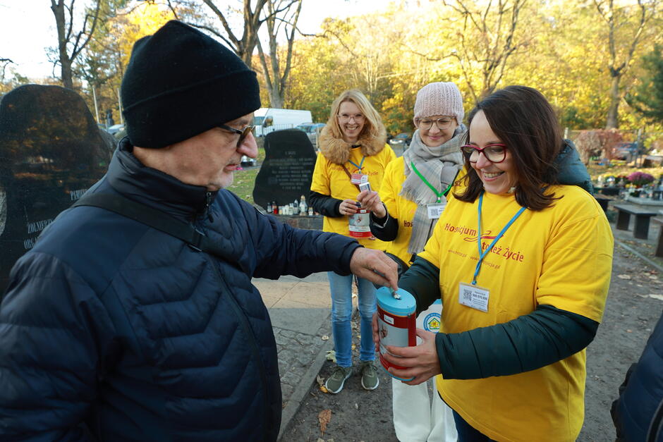 Dwie kobiety w żółtych koszulkach wolontariuszy zbierają datki do puszek na cmentarzu. Pieniądze wrzuca mężczyzna w czapce i okularach, obok stoi kobieta. Wszyscy się uśmiechają.