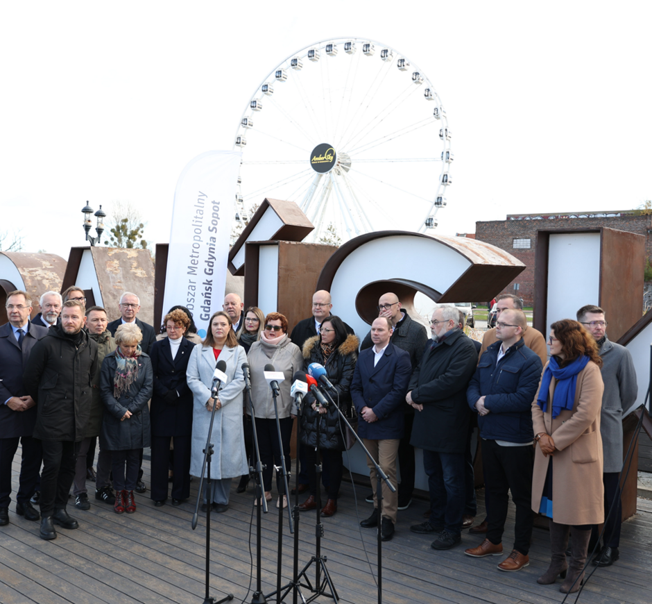 Na zdjęciu znajduje się grupa osób podczas oficjalnego wydarzenia, stojących przed dużym napisem "GDAŃSK", z widocznym diabelskim młynem w tle. Uczestnicy, w tym osoby przemawiające do mikrofonów, są zgromadzeni na drewnianym pomoście, a w tle widoczne są również flagi i budynki.