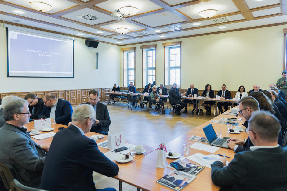 na zdjęciu duży stół ułożony w literę u, siedzi przy nim ponad dwadzieścia osób, wszyscy w oficjalnych strojach