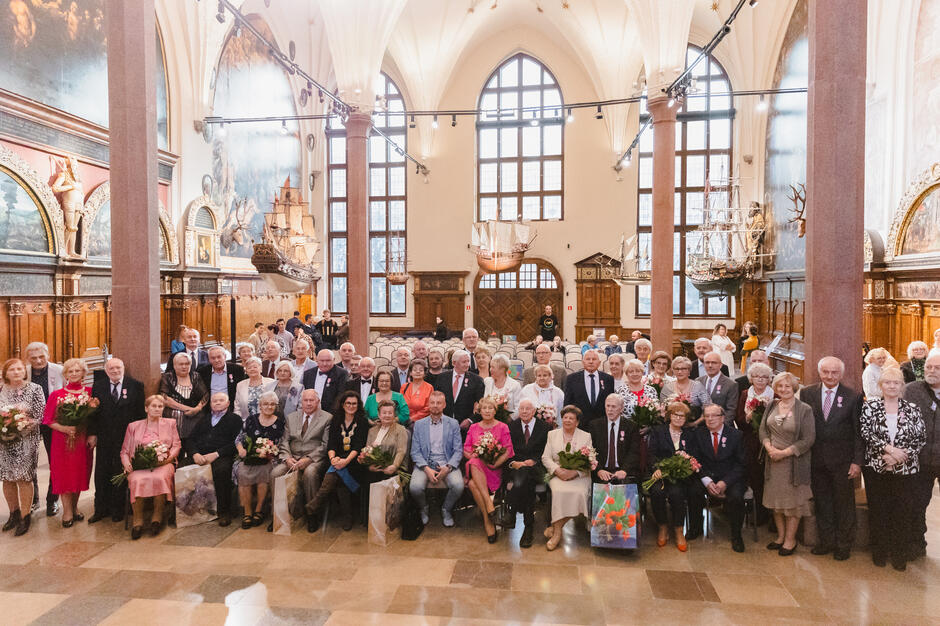 Zdjęcie przedstawia dużą grupę osób starszych, które pozują do pamiątkowej fotografii we wnętrzu eleganckiej sali z ozdobnymi elementami, modelami statków i freskami na ścianach