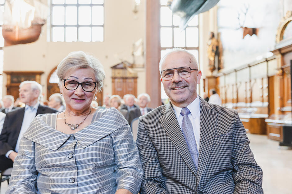 Na zdjęciu znajduje się uśmiechnięta para starszych osób, ubrana elegancko, siedząca w uroczystej sali z dużymi oknami i ozdobnym wystrojem. Kobieta ma na sobie stylową sukienkę w szare paski z ozdobnym kołnierzem, a mężczyzna jest w garniturze w kratę z krawatem