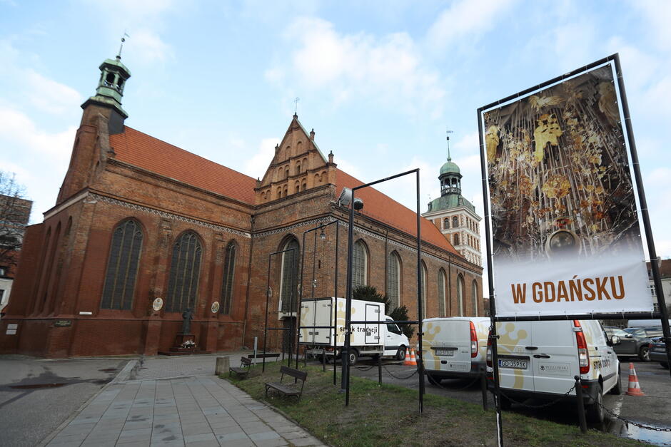 Zdjęcie przedstawia gotycki kościół z czerwonej cegły w Gdańsku, ozdobiony dużymi, wysokimi oknami oraz charakterystycznymi wieżyczkami. Na pierwszym planie widać plakat z napisem „W Gdańsku” i obrazem bursztynowego ołtarza, a obok zaparkowane są samochody dostawcze z lokalnym oznakowaniem