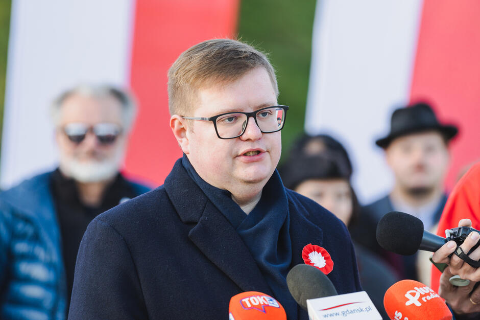 Na zdjęciu widzimy mężczyznę przemawiającego do mikrofonów kilku stacji radiowych i telewizyjnych. Ubrany jest w ciemny płaszcz, a na nim przypiętą ma biało-czerwoną kokardę – symbol patriotyczny, wskazujący na udział w wydarzeniu związanym z polskim świętem narodowym. Mężczyzna nosi okulary o prostokątnych oprawkach i ma krótkie, jasne włosy. W tle znajdują się rozmyte biało-czerwone flagi oraz inne osoby, które wyglądają na uczestników tego wydarzenia.