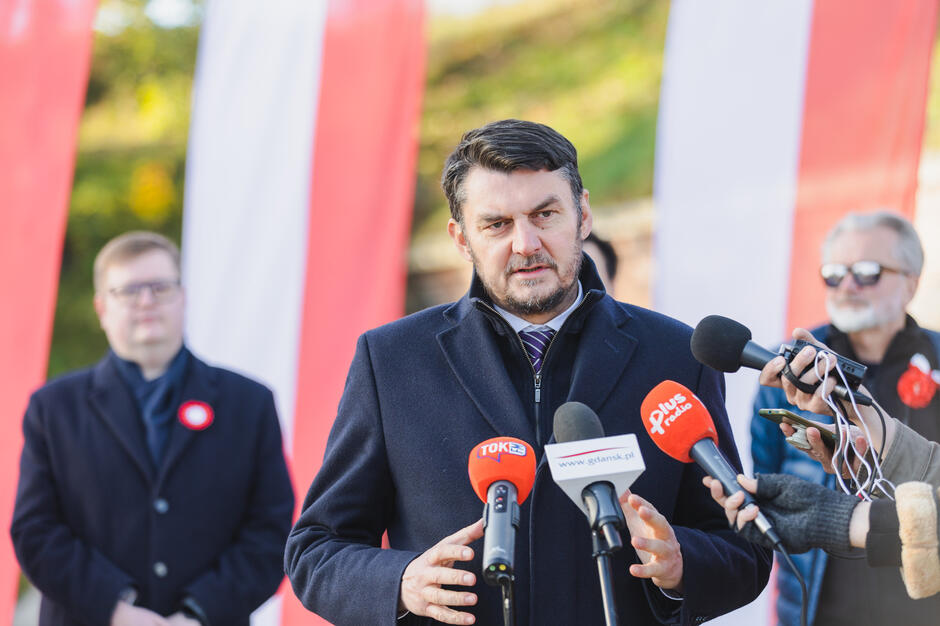 Na zdjęciu widzimy mężczyznę w ciemnym płaszczu przemawiającego do kilku mikrofonów różnych stacji radiowych i telewizyjnych, które są skierowane w jego stronę. Jest to oficjalne wydarzenie, o czym świadczy obecność biało-czerwonych flag w tle oraz kokarda patriotyczna na jego płaszczu. Mężczyzna ma krótko przystrzyżone ciemne włosy i brodę. Ubrany jest elegancko, a pod płaszczem widać krawat, co podkreśla formalny charakter wystąpienia. W tle stoją inne osoby, w tym mężczyzna w okularach przeciwsłonecznych, co sugeruje, że jest to publiczne wydarzenie z udziałem mediów.